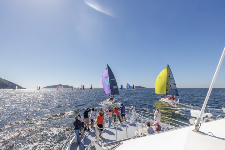 Sailboats racing on open water viewed from a nearby boat under a clear blue sky.