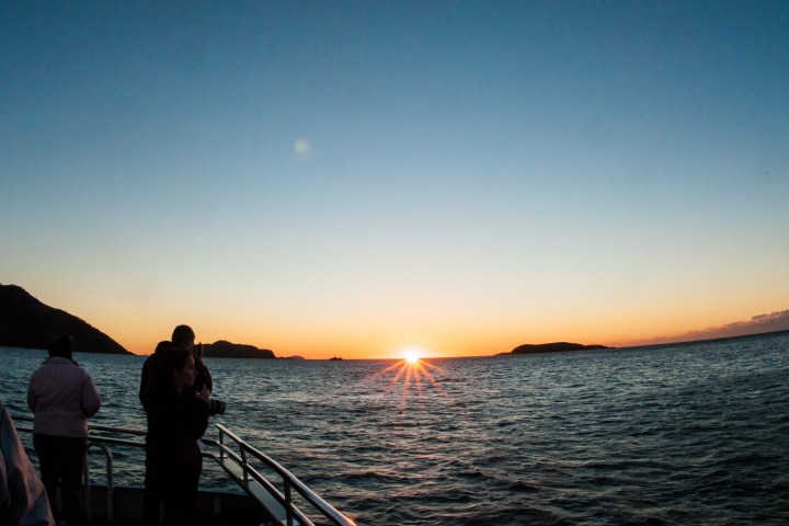People on a boat watch a sunset over the ocean with islands visible on the horizon.