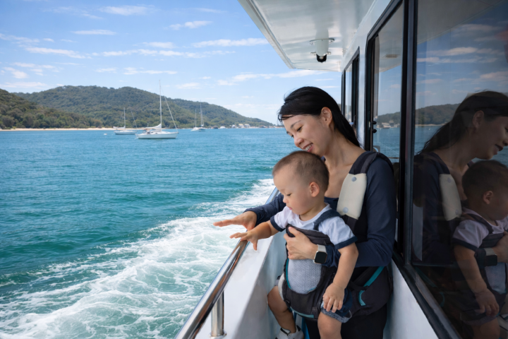 Woman with child on a boat, looking at the water, with a scenic coastline in the background.