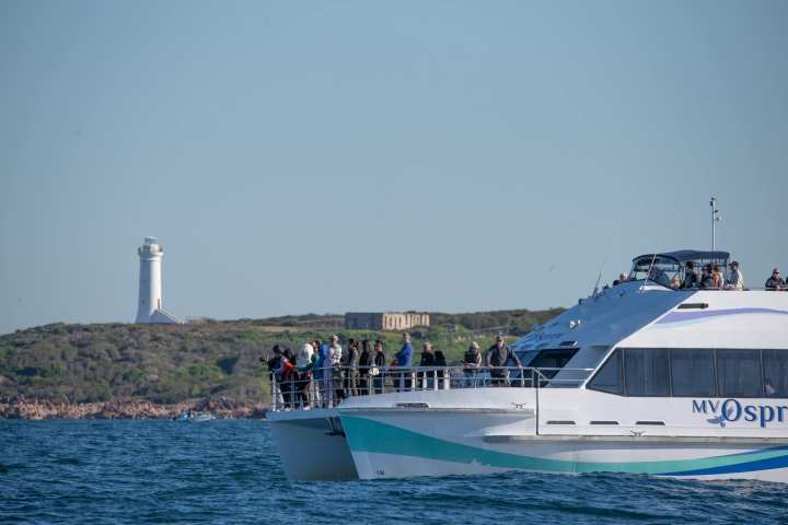 A ferry with passengers sails near a coastal lighthouse on a clear day.