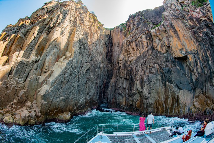 Boat approaching a tall rock formation with narrow gap over blue water.