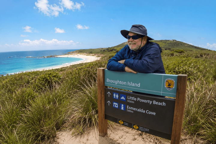Man in hat leaning on Broughton Island sign with beach and ocean in background.