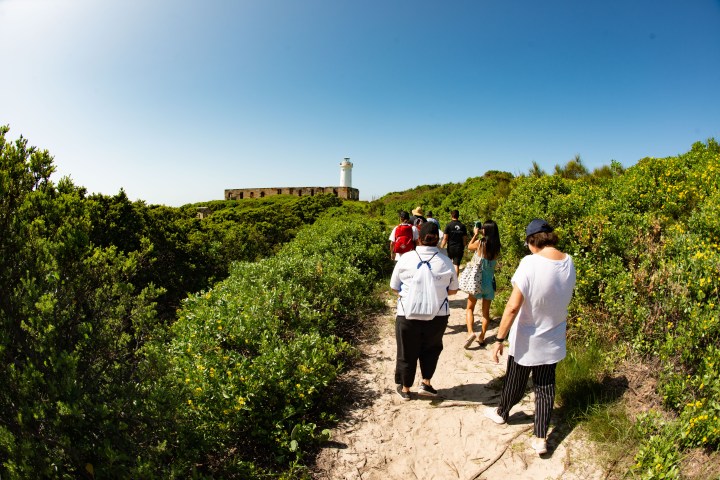 Group of people walking on a sandy path towards a lighthouse among green bushes under a clear blue sky.