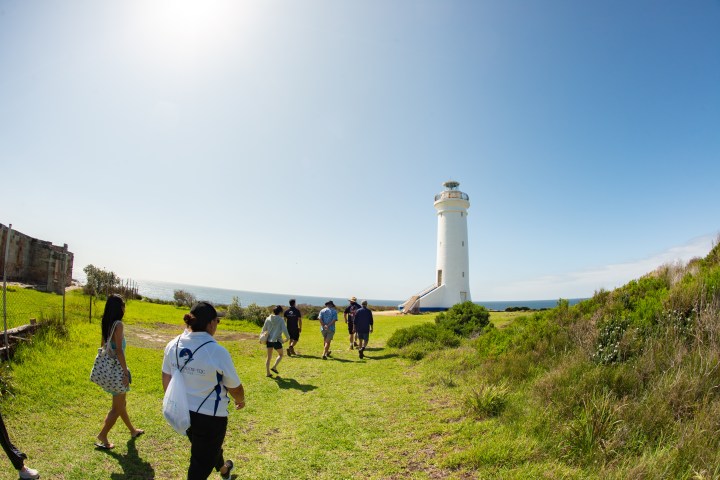 Group walking towards a white lighthouse on a grassy path by the sea.
