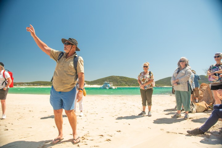 Group of people on a sunny beach, one person gesturing, with green water and hills in the background.