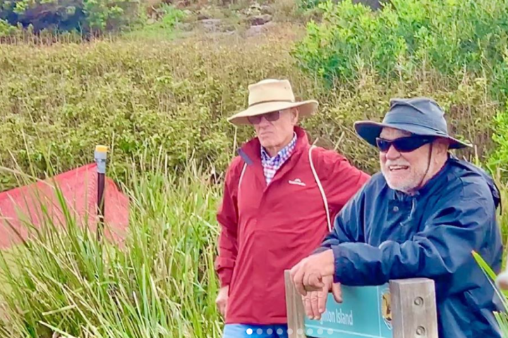 Two men in hats and jackets standing near a grassy area, one leaning on a wooden post.